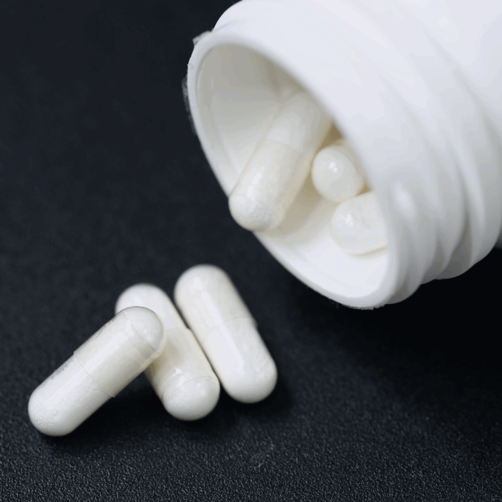A close-up photograph of white capsules spilling out of a white plastic container onto a dark surface.