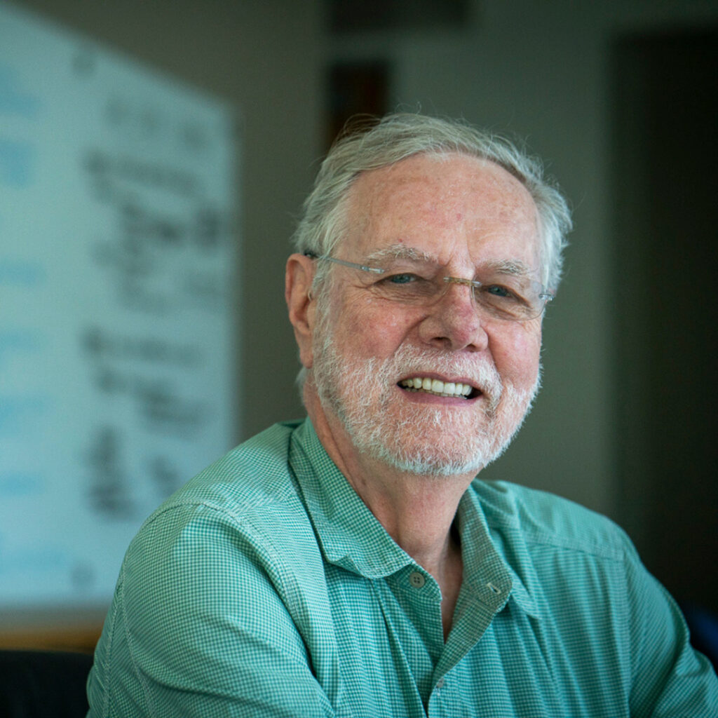 Ron Davis smiling, wearing glasses and a green button-down shirt, seated in an office with a blurred whiteboard in the background.